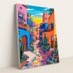 This artwork shows a vibrant, colorful street with blue, orange, and pink buildings, potted cacti, and a mosaic tile walkway, in a silver frame.