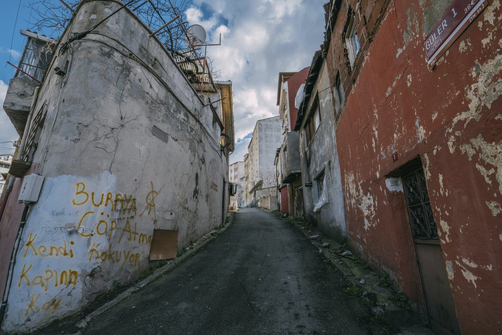 Sami TÜRK - A narrow, untidy urban alleyway featuring weathered buildings and graffiti under a cloudy sky.