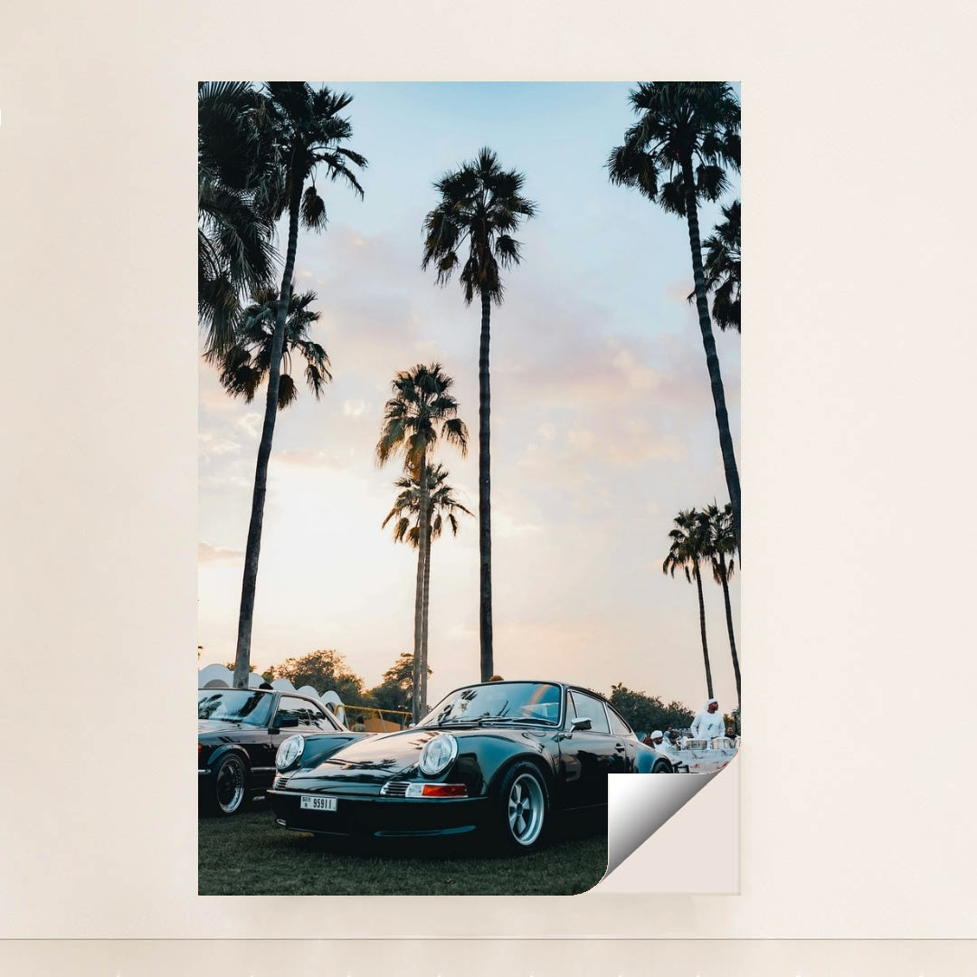 This photo shows a classic black Porsche sports car parked under tall palm trees at sunset, unframed.