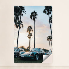 This photo shows a classic black Porsche sports car parked under tall palm trees at sunset, unframed.