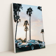 This photo shows a classic black Porsche sports car parked under tall palm trees at sunset, in a white frame.