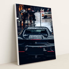 This photo shows the rear view of a black Ferrari covered in rain droplets on a wet city street at night, in a white frame.