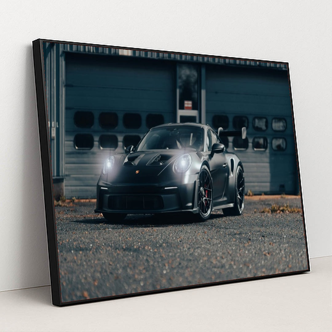This photo shows a black Porsche sports car with headlights on, parked on an industrial lot, in a black frame.