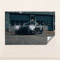 This photo shows a black Porsche sports car with headlights on, parked on an industrial lot, unframed.