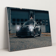 This photo shows a black Porsche sports car with headlights on, parked on an industrial lot, in a silver frame.