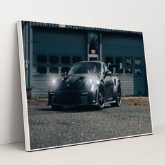 This photo shows a black Porsche sports car with headlights on, parked on an industrial lot, in a white frame.