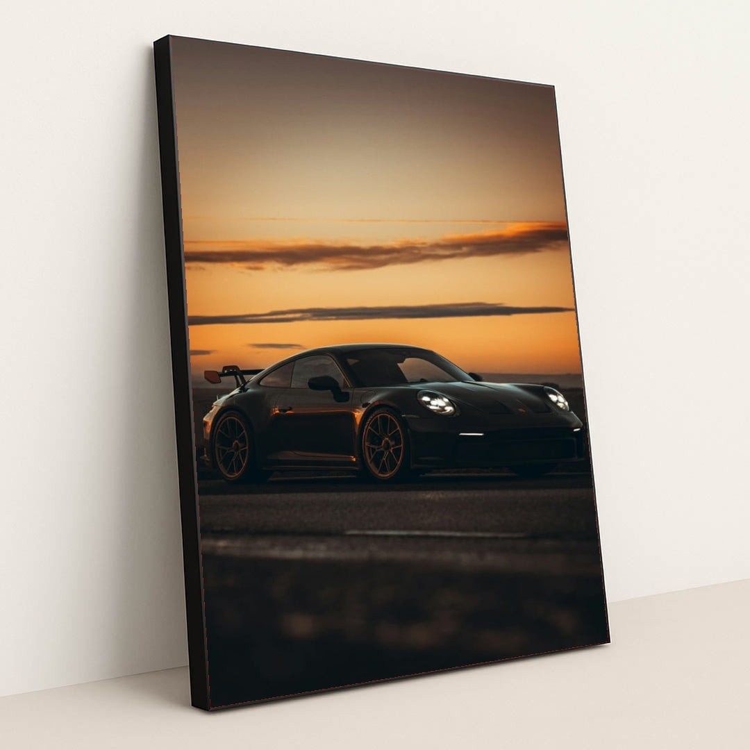 This photo shows a black Porsche sports car with a rear wing parked at sunset, in a black frame.