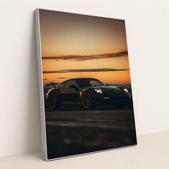 This photo shows a black Porsche sports car with a rear wing parked at sunset, in a silver frame.