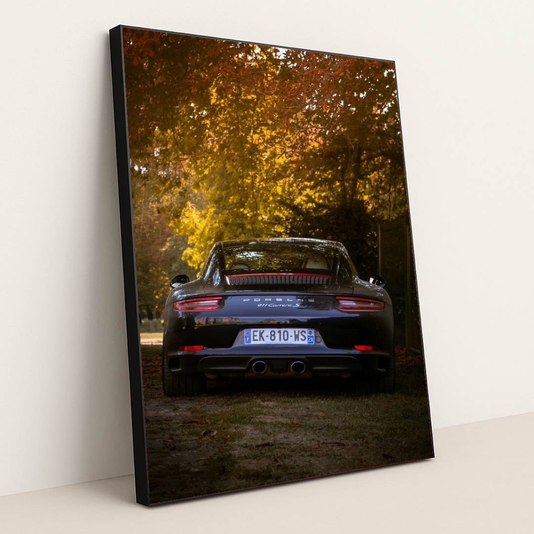 This photo shows the rear view of a black Porsche 911 Carrera S parked under autumn trees in soft evening light, in a black frame.