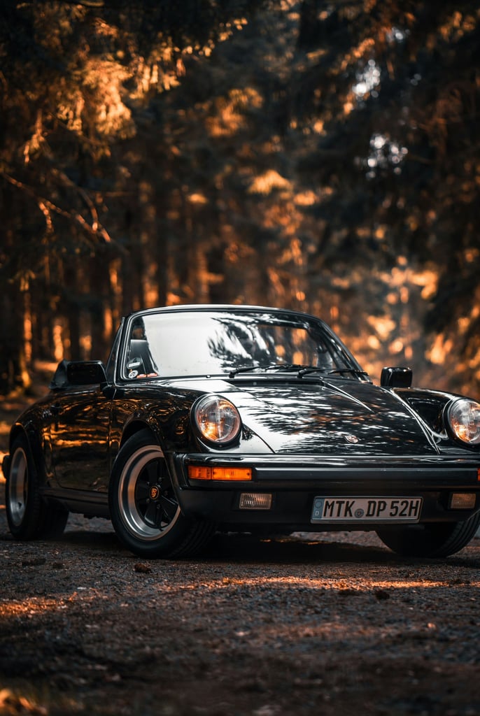Vintage Porsche 911 Forest Glow: black Cabriolet parked in a forest with tall trees and golden sunlight, showcasing classic design.