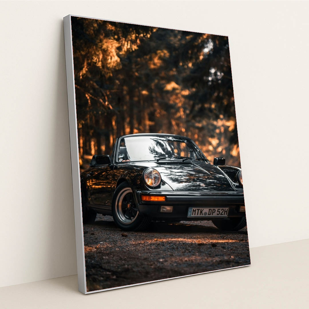 This photo shows a classic black Porsche convertible parked on a forest road with golden sunlight, in a silver frame.