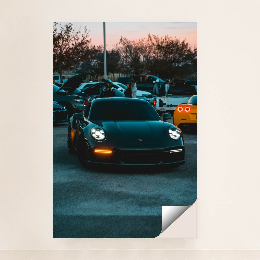 This photo shows a black Porsche 911 with LED headlights at dusk, parked at a car meet with people and vehicles in the background, unframed style.