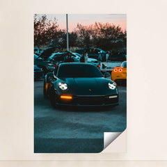 This photo shows a black Porsche 911 with LED headlights at dusk, parked at a car meet with people and vehicles in the background, unframed style.
