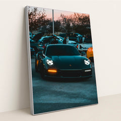 This photo shows a black Porsche 911 with LED headlights at dusk, parked at a car meet with people and vehicles in the background, in a silver frame.