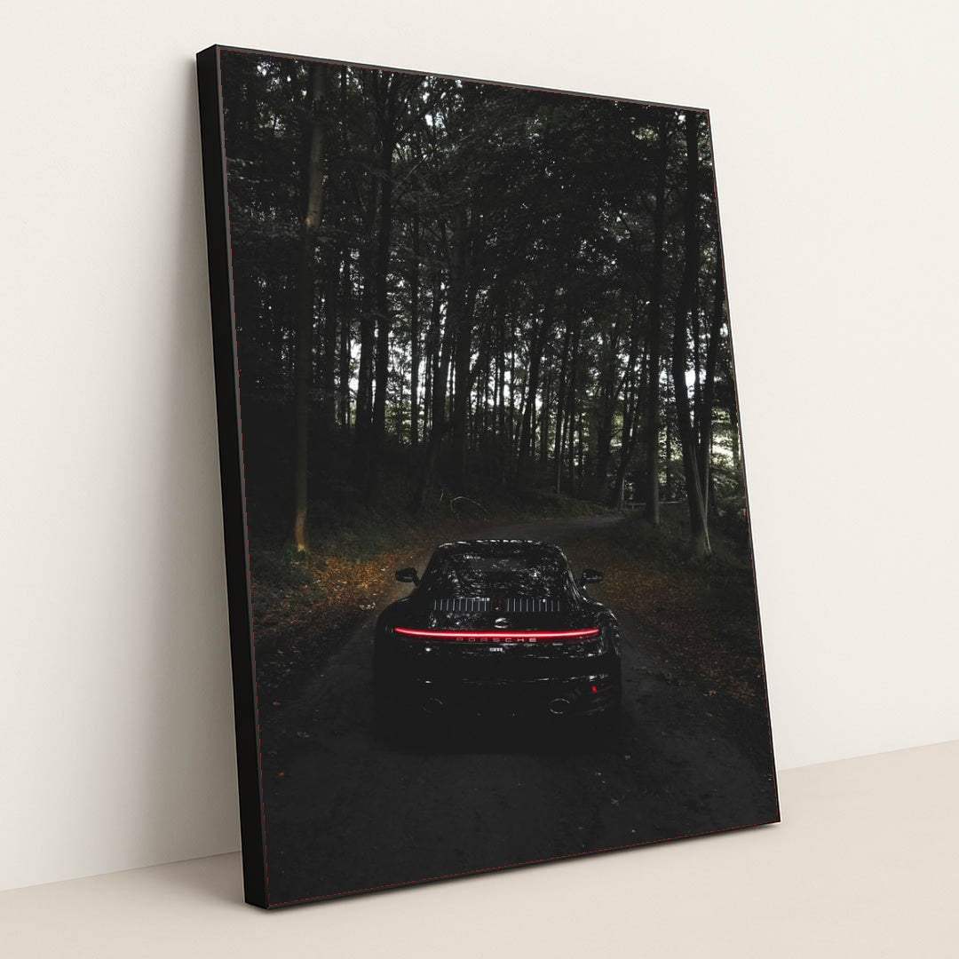This photo shows the rear of a black Porsche with taillights illuminated, parked on a dark forest road, in a black frame.