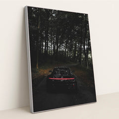 This photo shows the rear of a black Porsche with taillights illuminated, parked on a dark forest road, in a silver frame.