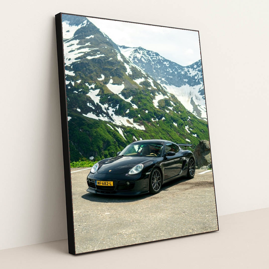 This photo shows a black Porsche sports car parked on a mountain road with snow-capped peaks in the background, in a black frame.