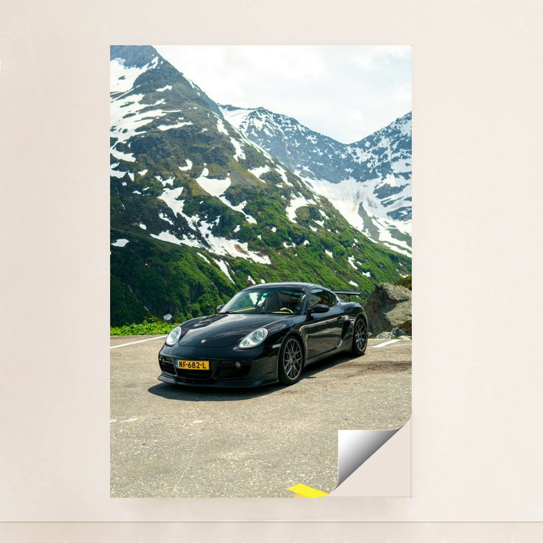 This photo shows a black Porsche sports car parked on a mountain road with snow-capped peaks in the background, unframed.
