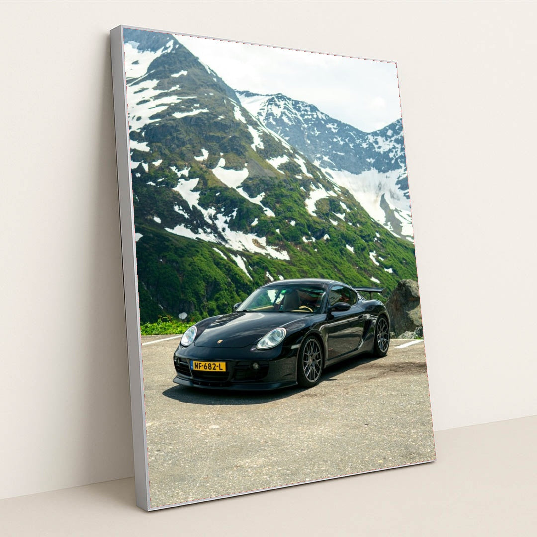 This photo shows a black Porsche sports car parked on a mountain road with snow-capped peaks in the background, in a silver frame.