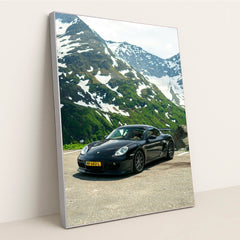 This photo shows a black Porsche sports car parked on a mountain road with snow-capped peaks in the background, in a silver frame.