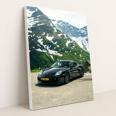 This photo shows a black Porsche sports car parked on a mountain road with snow-capped peaks in the background, in a white frame.