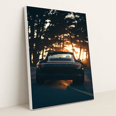 This photo shows the rear view of a black convertible Porsche parked on a tree-lined road at sunset, in a white frame.