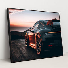 This photo shows a black Porsche sports car with a large rear wing parked at sunset near the ocean, in a black frame.