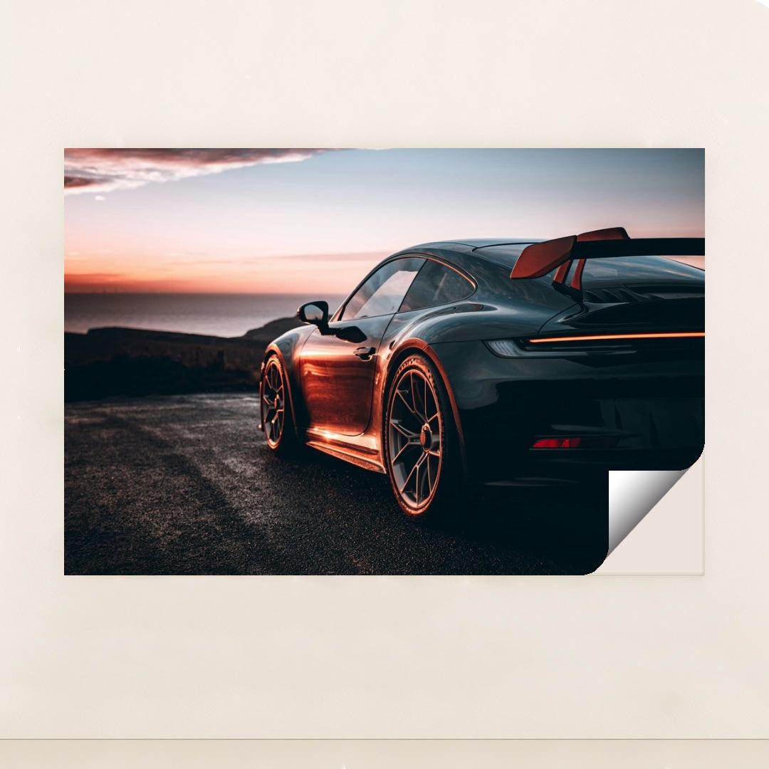 This photo shows a black Porsche sports car with a large rear wing parked at sunset near the ocean, unframed.