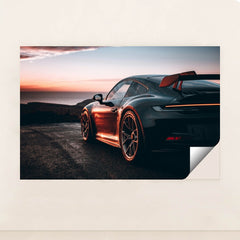 This photo shows a black Porsche sports car with a large rear wing parked at sunset near the ocean, unframed.