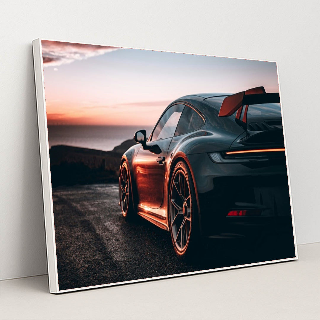 This photo shows a black Porsche sports car with a large rear wing parked at sunset near the ocean, in a white frame.