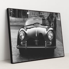 This photo shows a classic black Porsche Speedster convertible parked on a city street in black and white, in a black frame.