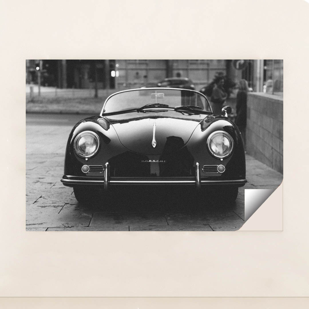 This photo shows a classic black Porsche Speedster convertible parked on a city street in black and white, minimalist unframed style.