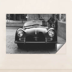 This photo shows a classic black Porsche Speedster convertible parked on a city street in black and white, minimalist unframed style.