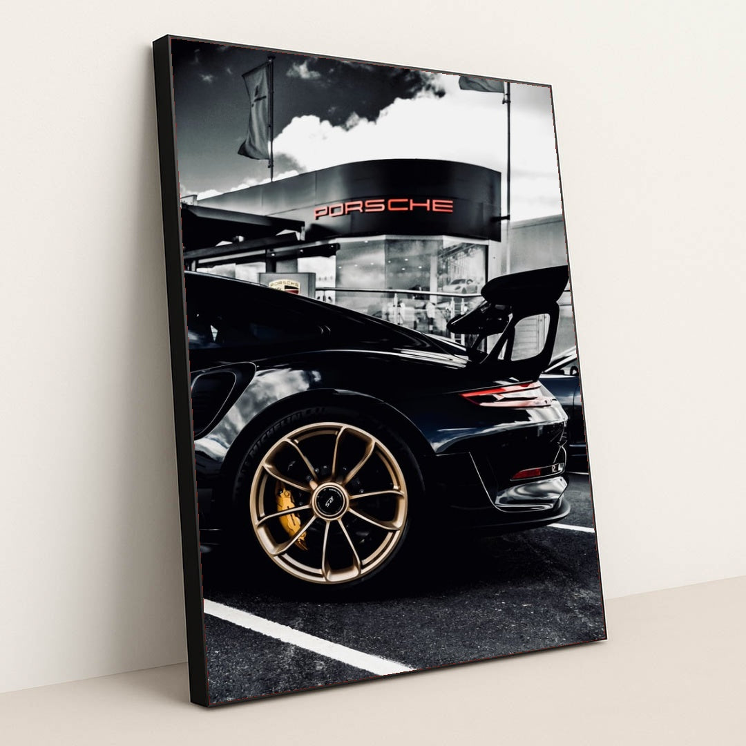 This photo shows the rear side view of a black Porsche GT sports car with gold wheels parked outside a dealership, in a black frame.