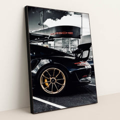 This photo shows the rear side view of a black Porsche GT sports car with gold wheels parked outside a dealership, in a black frame.