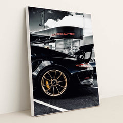 This photo shows the rear side view of a black Porsche GT sports car with gold wheels parked outside a dealership, in a white frame.