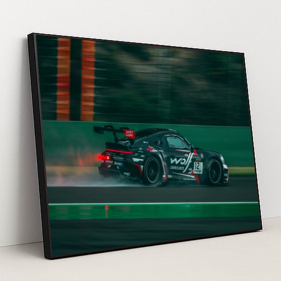 This photo shows a black Porsche race car with sponsor decals driving fast on a wet racetrack, in a black frame.