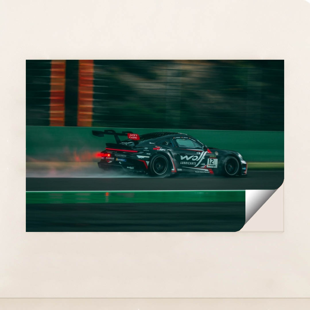 This photo shows a black Porsche race car with sponsor decals driving fast on a wet racetrack, unframed style.