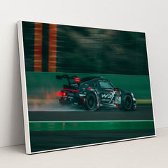 This photo shows a black Porsche race car with sponsor decals driving fast on a wet racetrack, in a white frame.