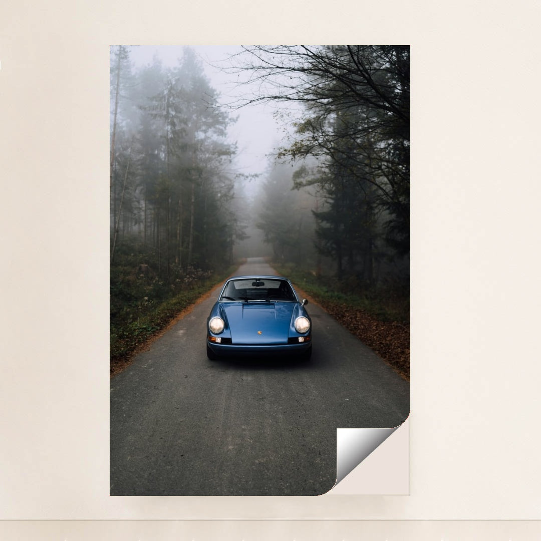 This photo shows a classic blue Porsche 911 parked on an empty forest road in morning mist, minimalist unframed style.