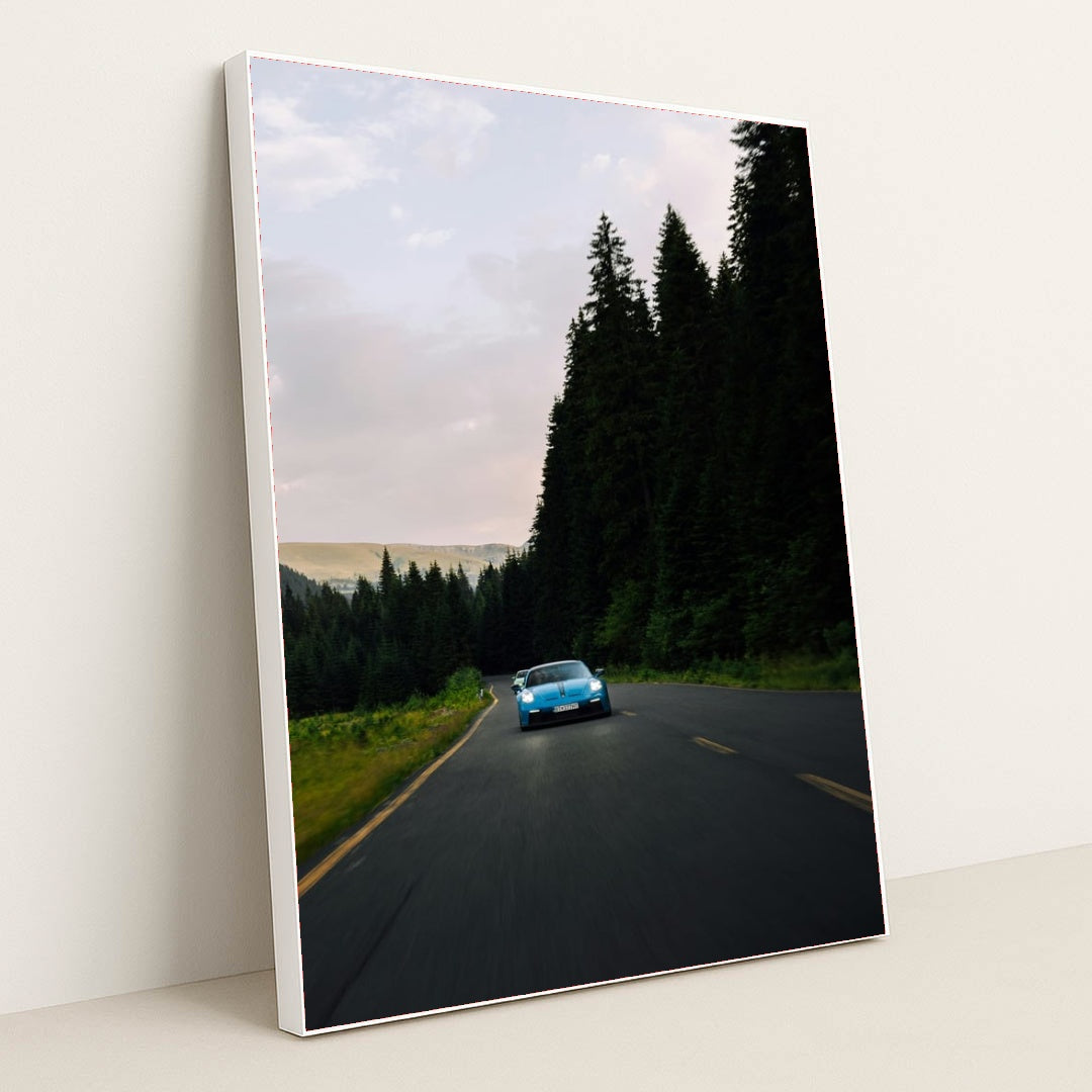 This photo shows a blue Porsche sports car driving on a curvy forest road at dusk, in a white frame.