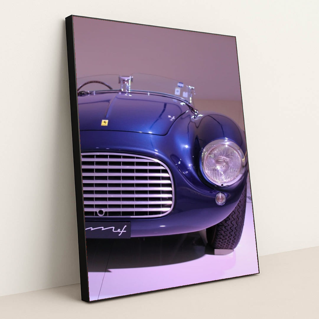 This photo shows the front left side of a blue vintage Ferrari convertible under display lights, in a black frame.