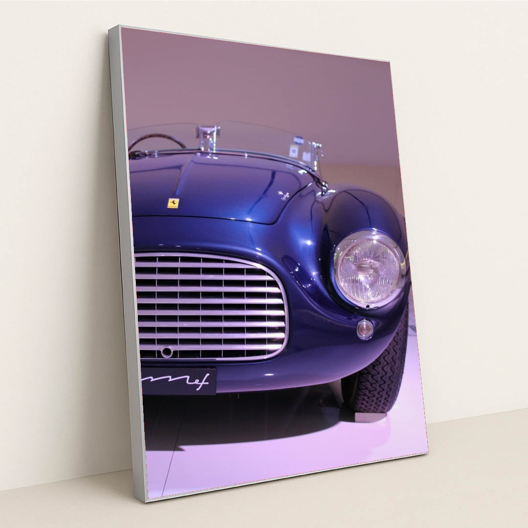 This photo shows the front left side of a blue vintage Ferrari convertible under display lights, in a silver frame.