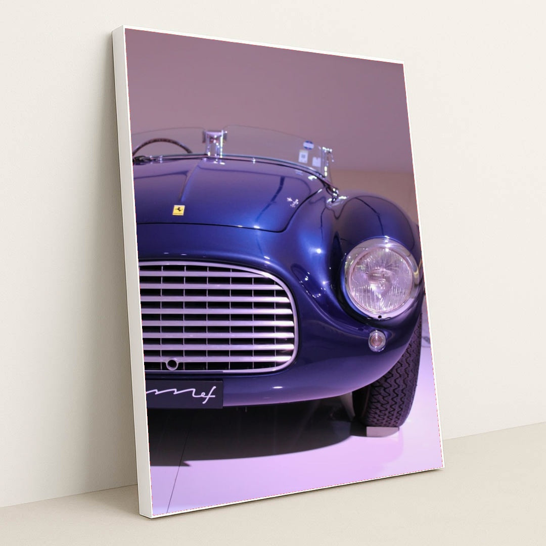 This photo shows the front left side of a blue vintage Ferrari convertible under display lights, in a white frame.