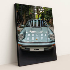 This photo shows a pastel blue classic Porsche 911 coupe on display in a modern indoor courtyard with trees, in a black frame.
