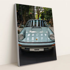 This photo shows a pastel blue classic Porsche 911 coupe on display in a modern indoor courtyard with trees, in a silver frame.