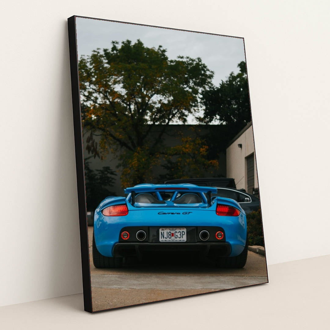This photo shows a bright blue Porsche Carrera GT viewed from the rear, parked outside with trees and buildings in the background, in a black frame.