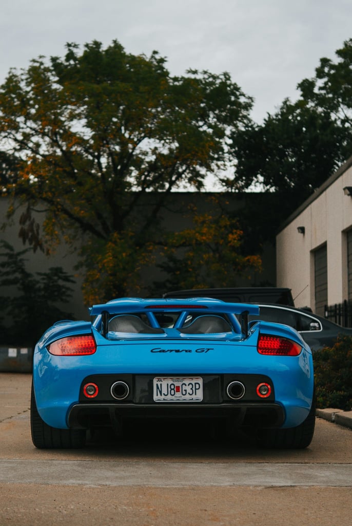 Porsche Carrera GT in Blue Steel Silence showcasing the sleek rear and carbon wing against an industrial backdrop.