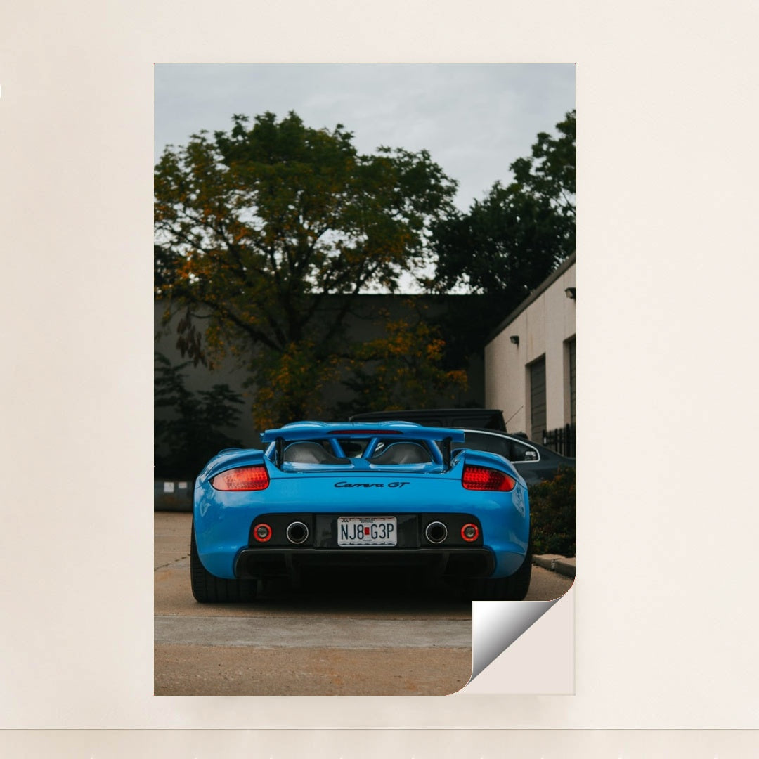 This photo shows a bright blue Porsche Carrera GT viewed from the rear, parked outside with trees and buildings in the background, unframed style.