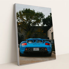 This photo shows a bright blue Porsche Carrera GT viewed from the rear, parked outside with trees and buildings in the background, in a silver frame.